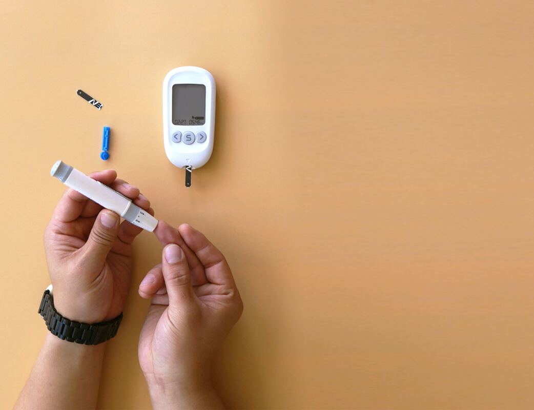 A pair of hands use a lancing device to prick a finger and test for diabetes with a Blood Glucose Meter against a yellow-tan backdrop.