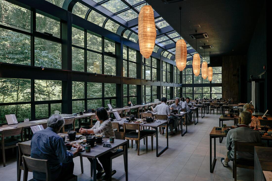 Diners sit in a restaurant with tables and chairs, paper lantern light fixtures, and many windows.