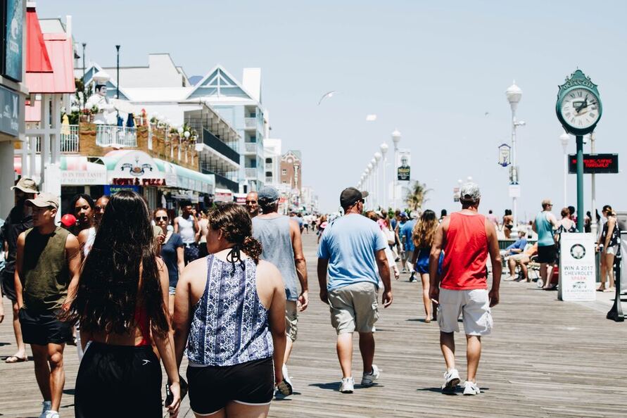 A man with hearing aids, a baseball hat, a light blue t-shirt, and khaki shorts walks on the boardwalk in a summer heatwave.