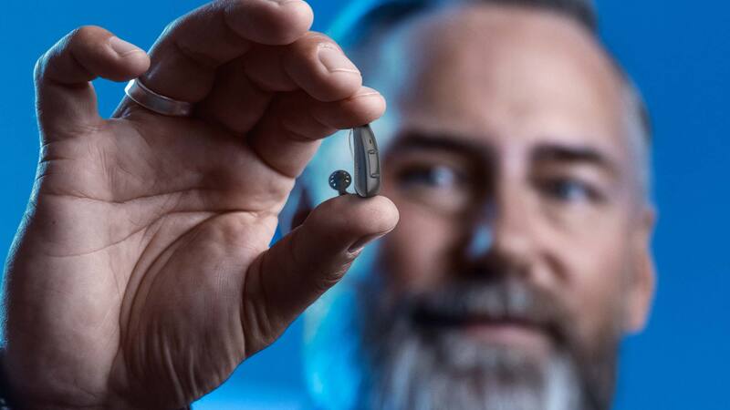 bearded man holds silver behind-the-ear hearing aid with receiver